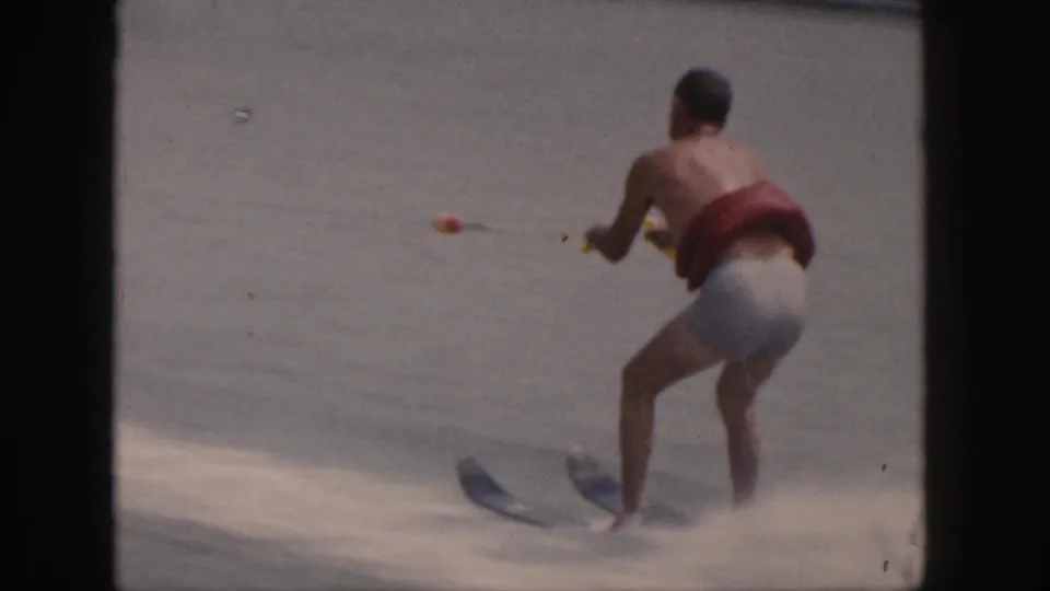 A man water skis on a Michigan lake in 1961, wearing swim trunks and a red life vest while gripping yellow tow ropes.
