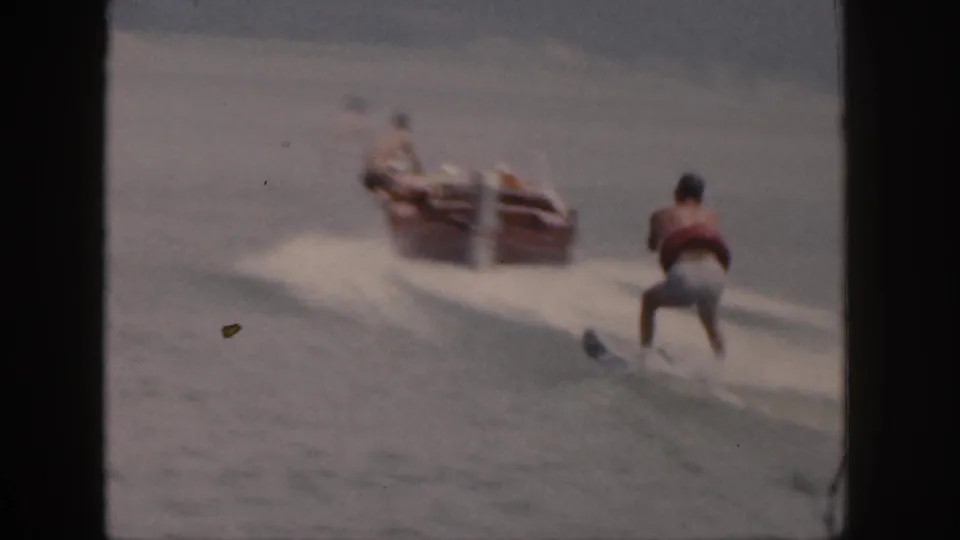 The skier trails behind a small motorboat on a Michigan lake, with water spray and soft 8mm blur across the frame.