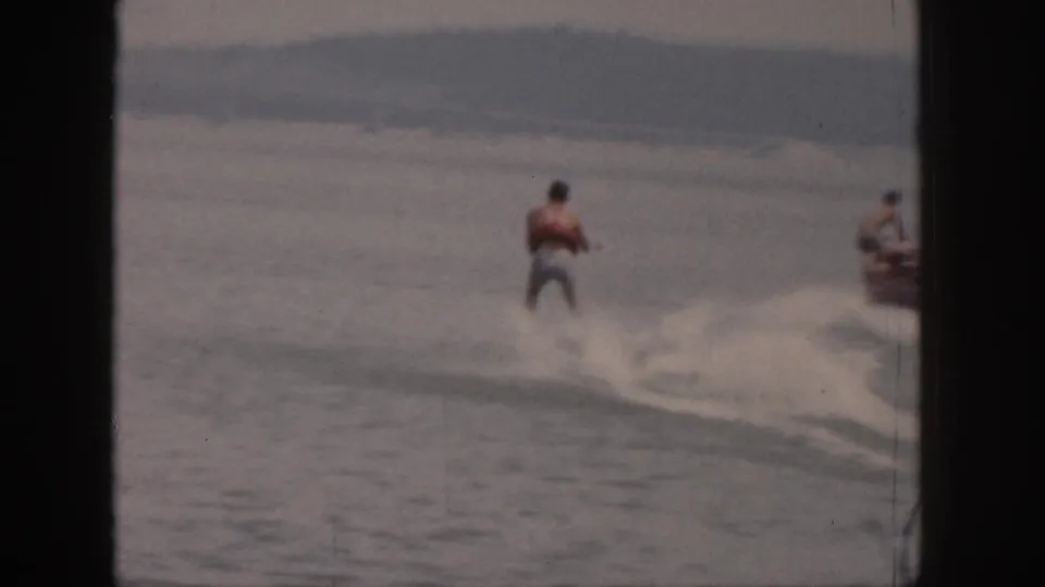 A 1961 Michigan lake view shows the skier moving away through spray, with a boat at the edge and hills or shoreline in the distance.
