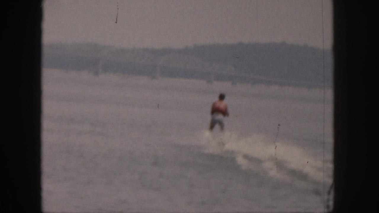 Distant water skier in a red vest moving across a gray lake.