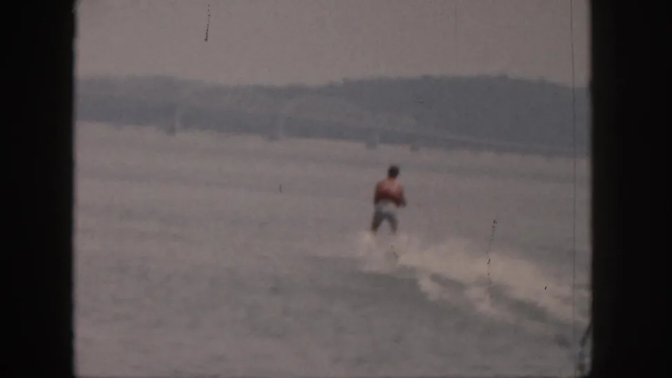 1961 Michigan home-movie frame showing a distant adult water skier in a red vest crossing the lake surface.