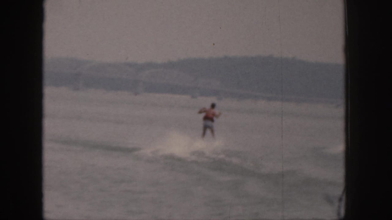 A distant adult water skier in a red vest rides over a low wake on a Michigan lake in 1961 footage.
