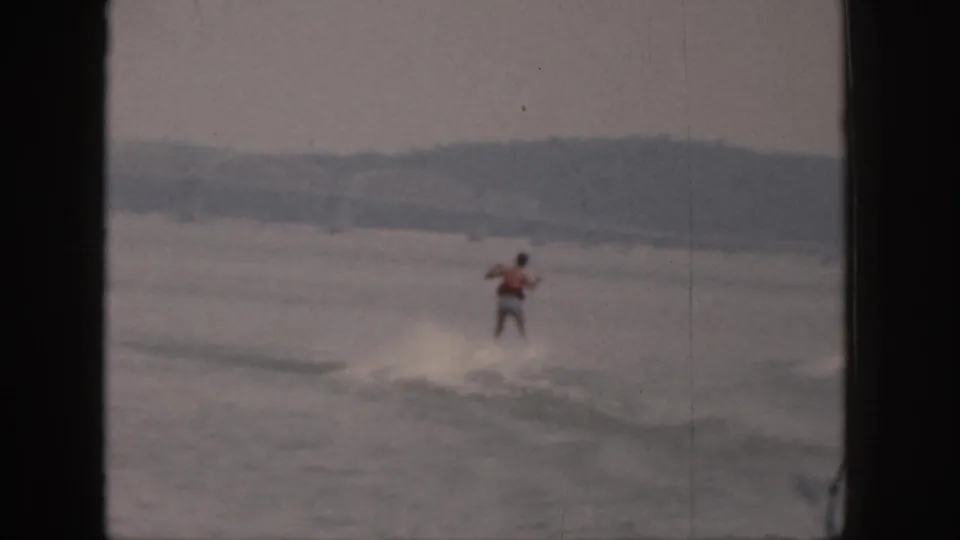 A distant adult water skier in a red vest rides over a low wake on a Michigan lake in 1961 footage.