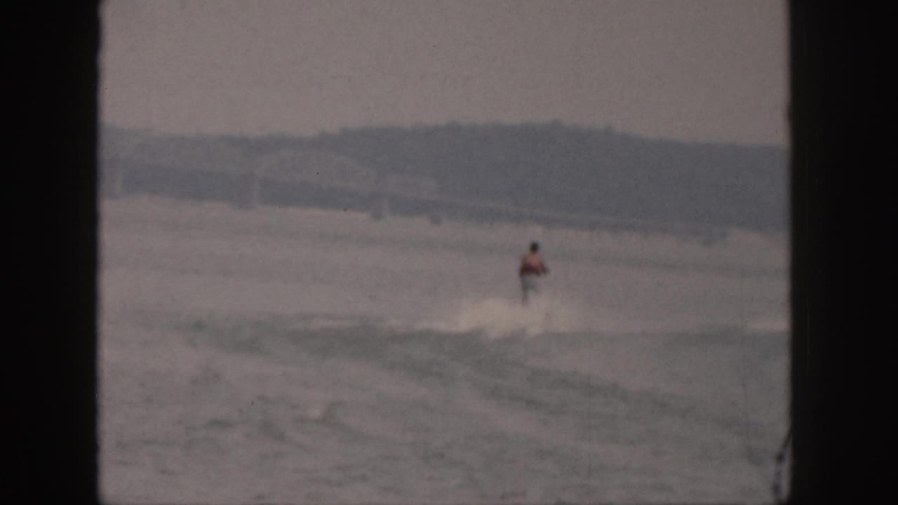 The home-movie camera holds a distant view of the skier continuing across the lake with a low shoreline beyond.