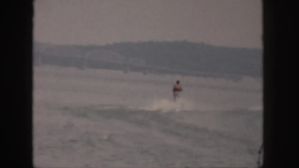 The home-movie camera holds a distant view of the skier continuing across the lake with a low shoreline beyond.
