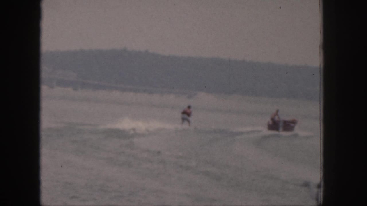A distant skier moves near the wake while a small motorboat appears at the right edge of the 1961 Michigan lake scene.