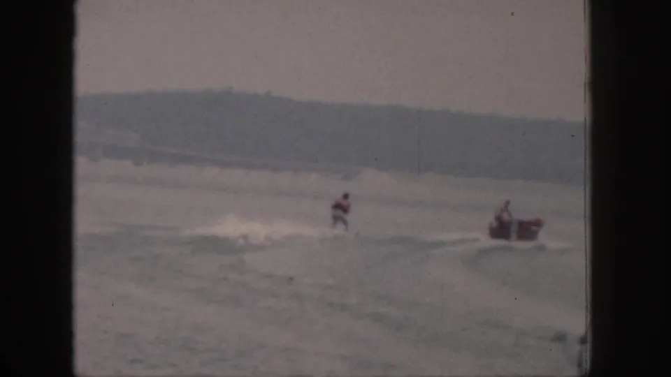 A distant skier moves near the wake while a small motorboat appears at the right edge of the 1961 Michigan lake scene.
