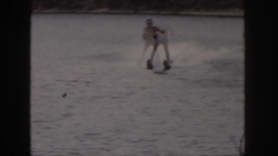 1961 Michigan home-movie frame showing a distant man upright on water skis with the shoreline faintly behind him.