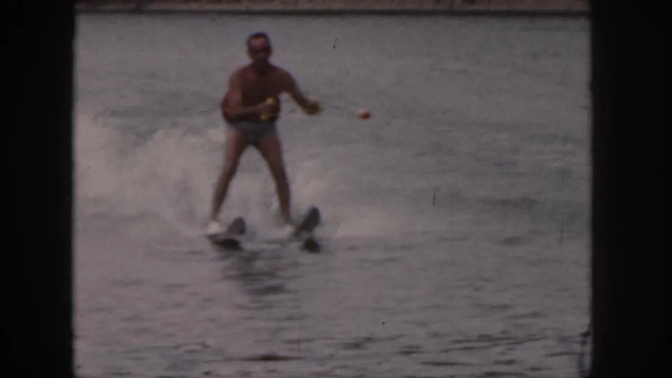 1961 Michigan 8mm frame of a man in a red life vest and light trunks balancing on water skis near the camera.