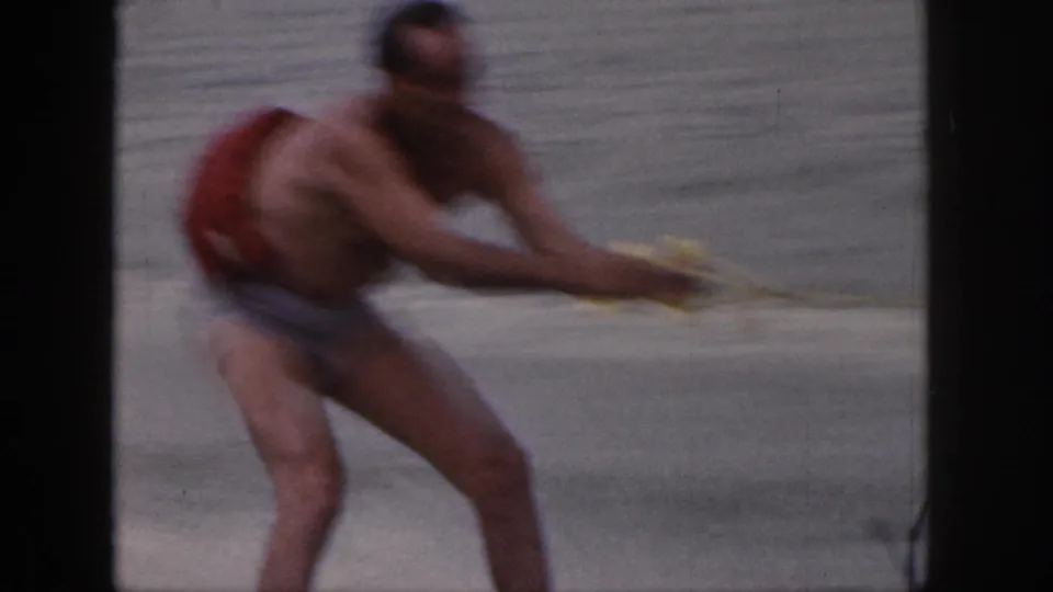 Close 1961 Michigan 8mm frame of the waterskier leaning with a yellow tow handle, red life vest, and lake water behind him.