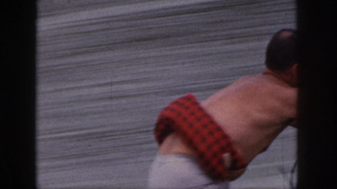 Shirtless man in red plaid trunks leans forward at the edge of a waterskiing shot.