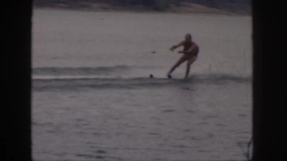 A distant 1961 Michigan home-movie frame shows the man balancing on waterskis over a low wake.