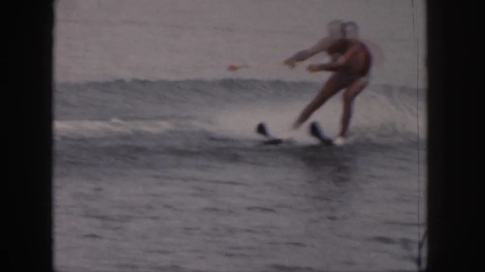The older man in red trunks leans through a turn on waterskis, framed by lake spray and Super 8 gate edges.