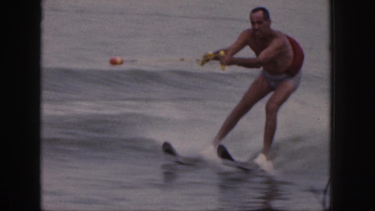 Man in swim trunks and red life vest water skiing on a calm lake.