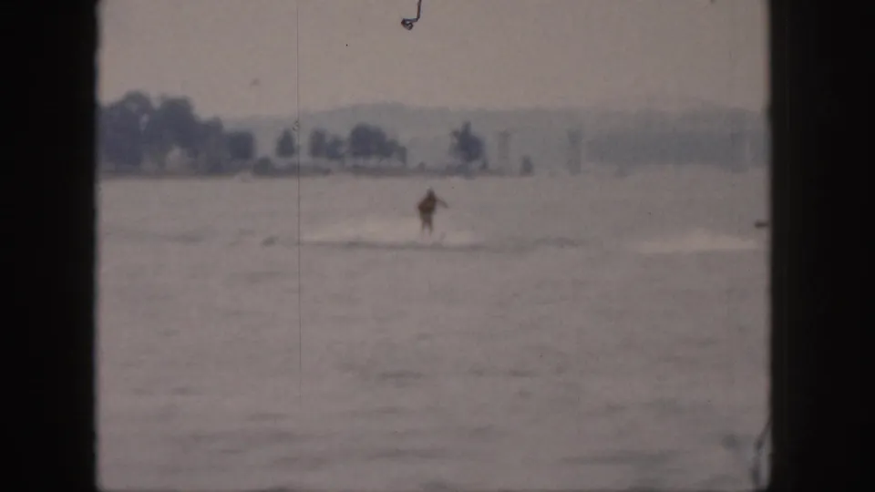 Wide 1961 Michigan lake view with the water skier distant on the water and a tree-lined shore beyond.