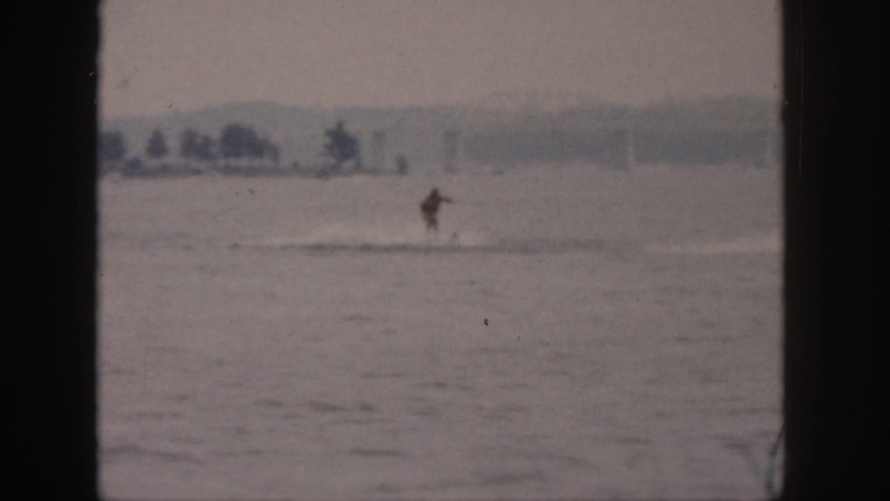 1961 Michigan 8mm frame showing a distant person water skiing across a calm river with a hazy shoreline beyond.