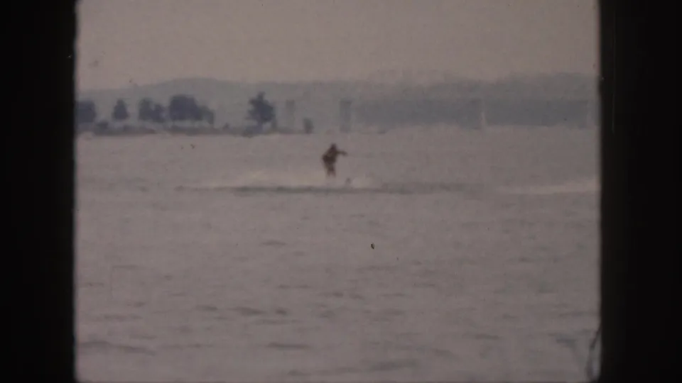 1961 Michigan 8mm frame showing a distant person water skiing across a calm river with a hazy shoreline beyond.