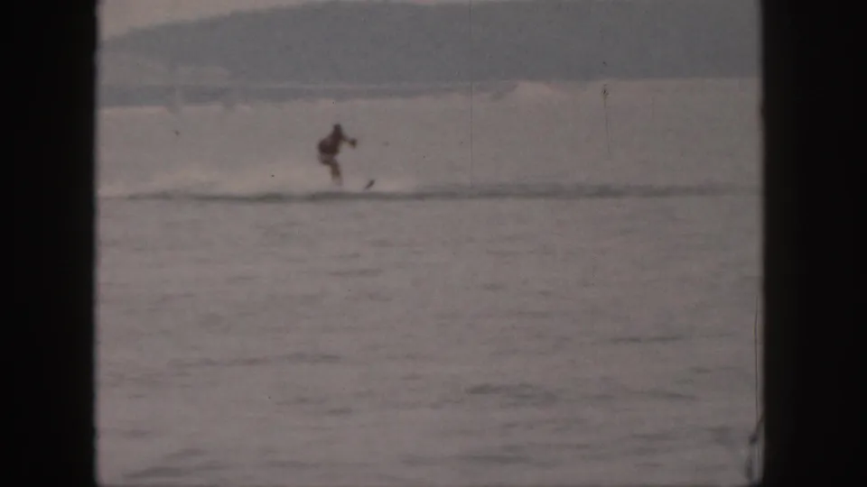 1961 Michigan home-movie still of a person water skiing at distance on a calm river, with soft grain and faded color.