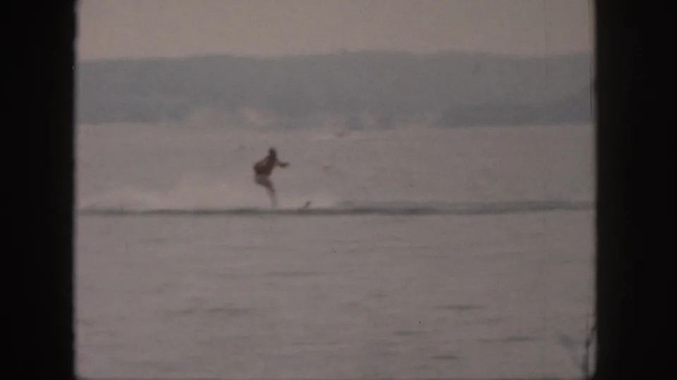 Final 1961 Michigan frame showing a small water skier moving along a river, with a pale wake and wooded shore in the distance.