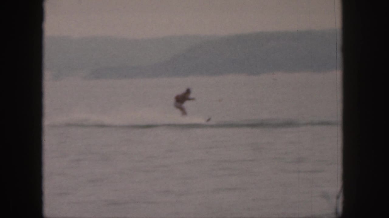 1961 Michigan 8mm frame showing a distant male water skier crossing a calm lake with wooded shoreline beyond.