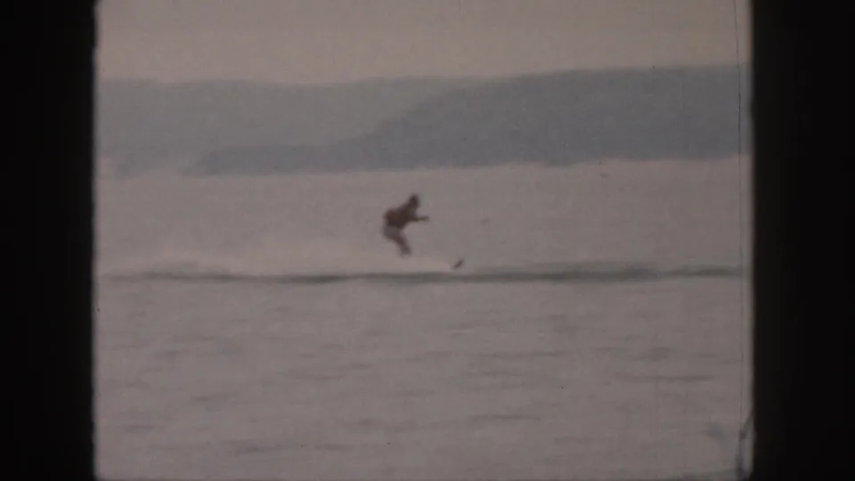 1961 Michigan 8mm frame showing a distant male water skier crossing a calm lake with wooded shoreline beyond.