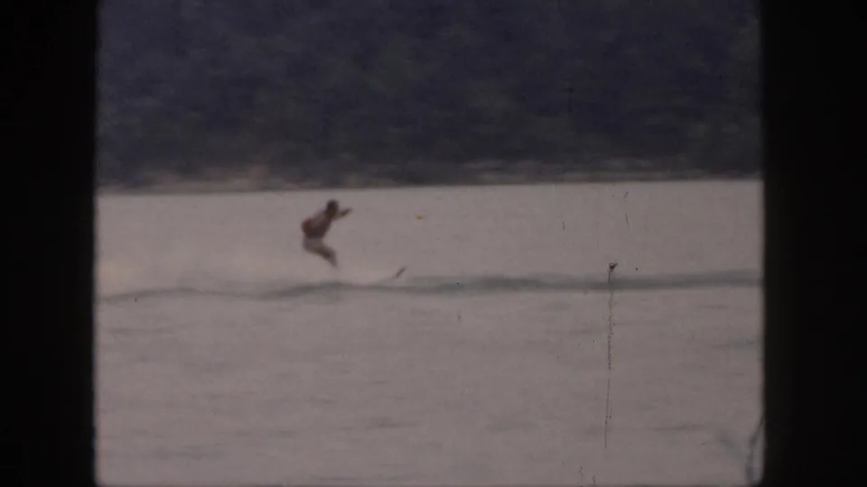 Distant 1961 Michigan lake view with a male water skier leaning forward as wake trails behind him.
