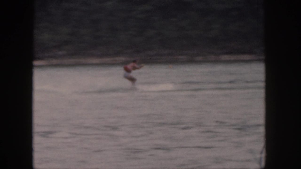 1961 Michigan home-movie frame showing a distant man water-skiing across a lake, with trees along the far shore.