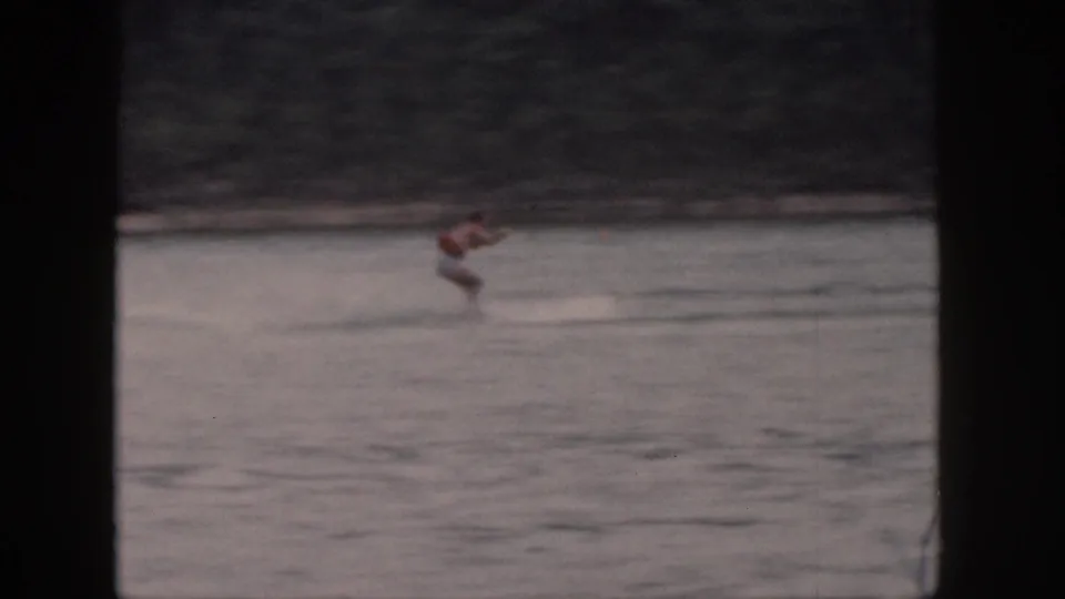 1961 Michigan home-movie frame showing a distant man water-skiing across a lake, with trees along the far shore.