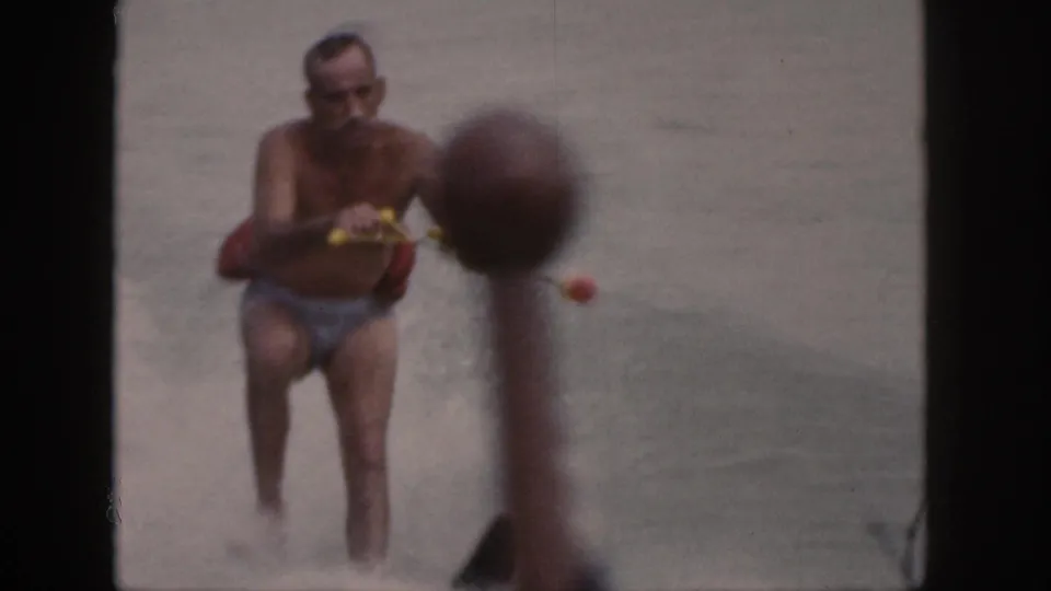 A man in swim trunks holds the tow rope in the lake after the fall, with water spray and shoreline visible behind him.