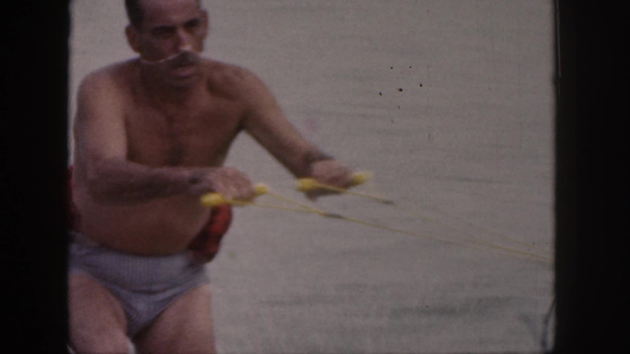1961 Michigan 8mm frame of a shirtless adult man holding a yellow tow rope while attempting to water ski.
