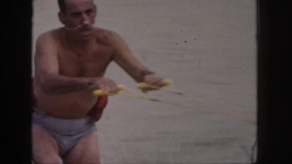 1961 Michigan 8mm frame of a shirtless adult man holding a yellow tow rope while attempting to water ski.