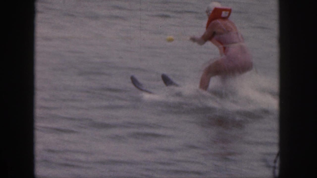 Woman in a red life vest skis across the lake, leaving spray behind her.