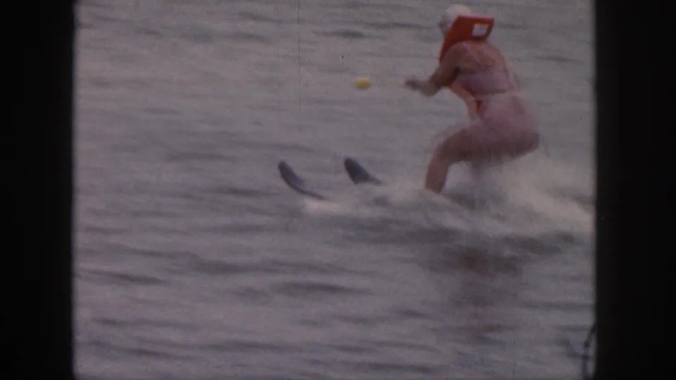 1961 Michigan home-movie frame showing an adult woman in a red life vest water skiing across a lake.