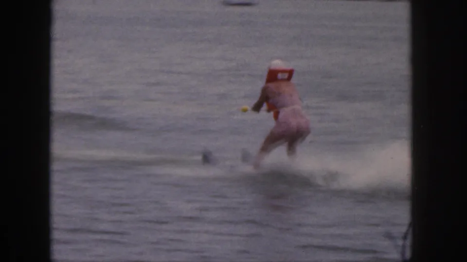 Adult woman in a red life vest water skis away from the camera in 1961 Michigan lake footage.