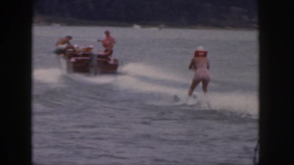 1961 Michigan 8mm frame with a small motorboat towing an adult woman water skier across open water.