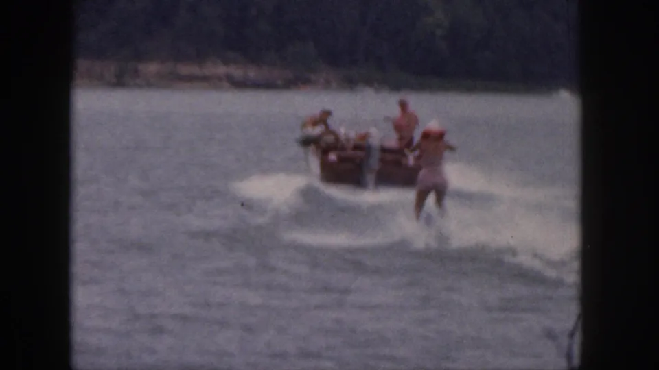 1961 Michigan 8mm home-movie frame showing a motorboat pulling a water skier in red across a calm lake.
