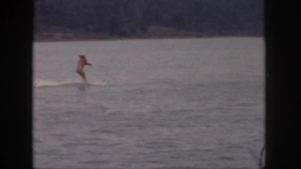 1961 Michigan home-movie frame showing a distant skier in red crossing the lake after the tow run continues.