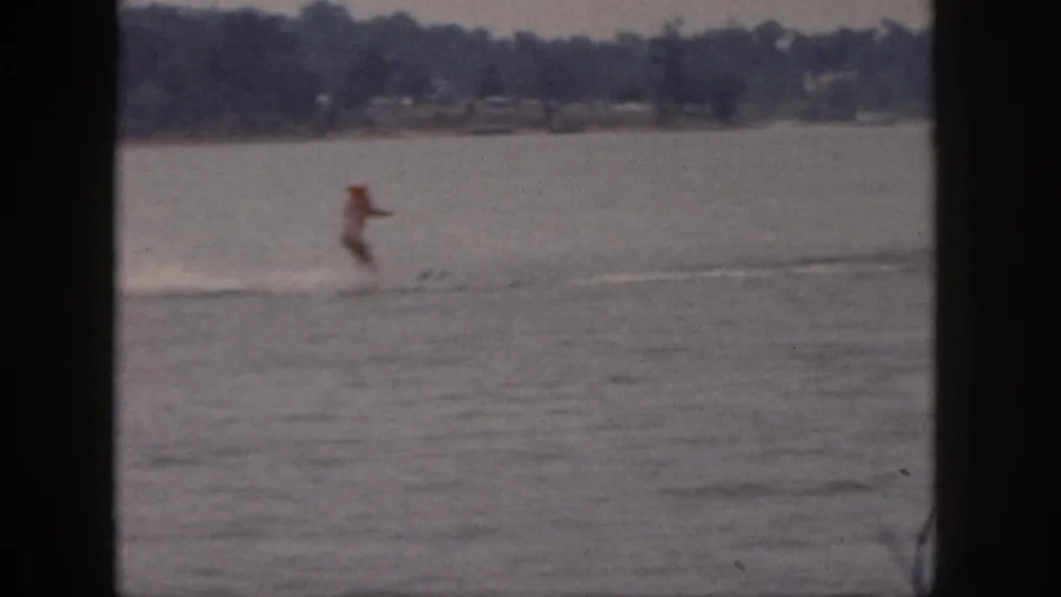 1961 Michigan 8mm frame of the skier moving across open lake water, with distant shore and homes faintly visible.