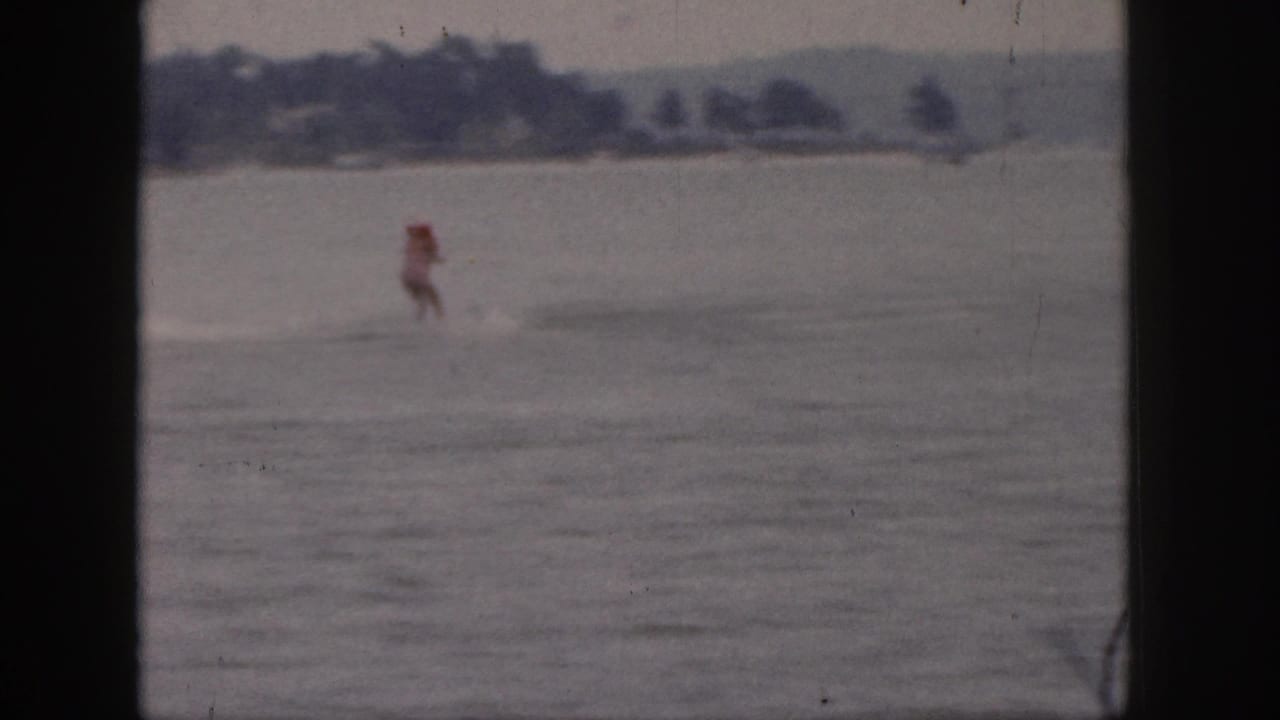 1961 Michigan home-movie frame showing a distant red-topped rider crossing a calm lake with a low wooded shore behind.