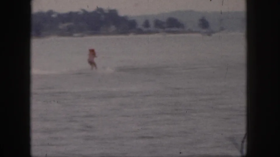 1961 Michigan home-movie frame showing a distant red-topped rider crossing a calm lake with a low wooded shore behind.