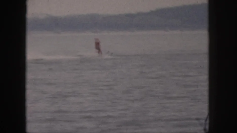 Distant 1961 Michigan lake recreation frame with the red-topped rider moving over the water and shoreline in the background.
