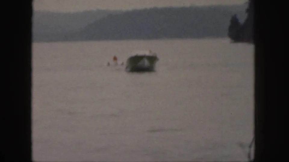 1961 Michigan home-movie frame showing a small motorboat on the lake with distant figures and a tree-lined shore in view.