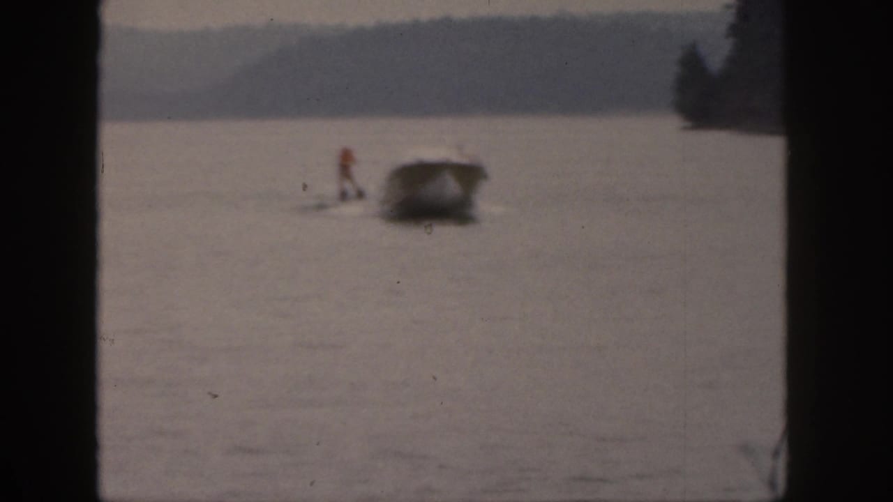 A distant motorboat pulls a water skier across a lake near a wooded shoreline.
