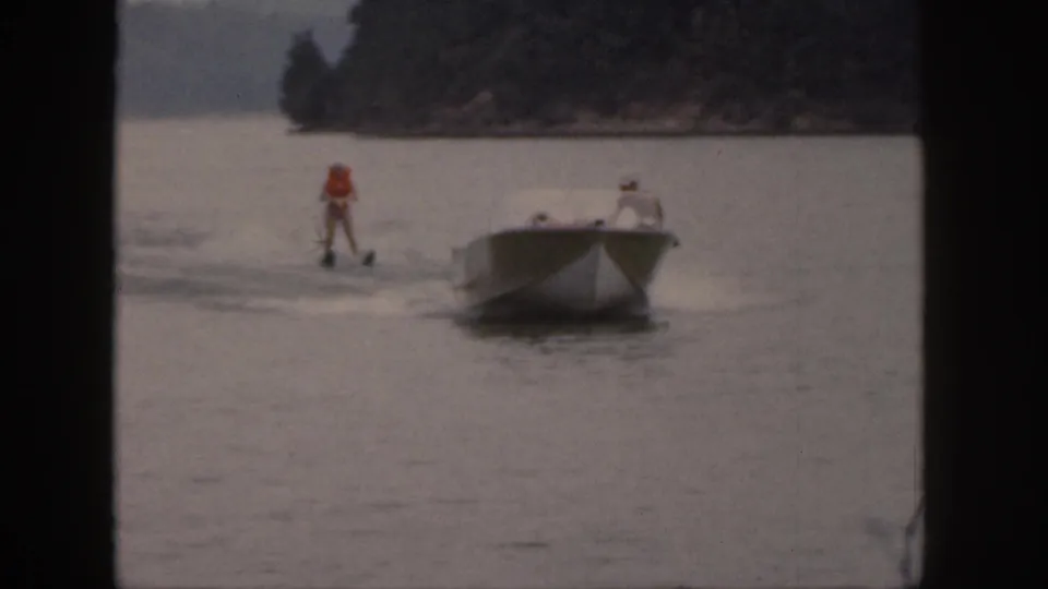In 1961 Michigan home-movie footage, a skier in an orange life vest follows a white motorboat across a calm lake.