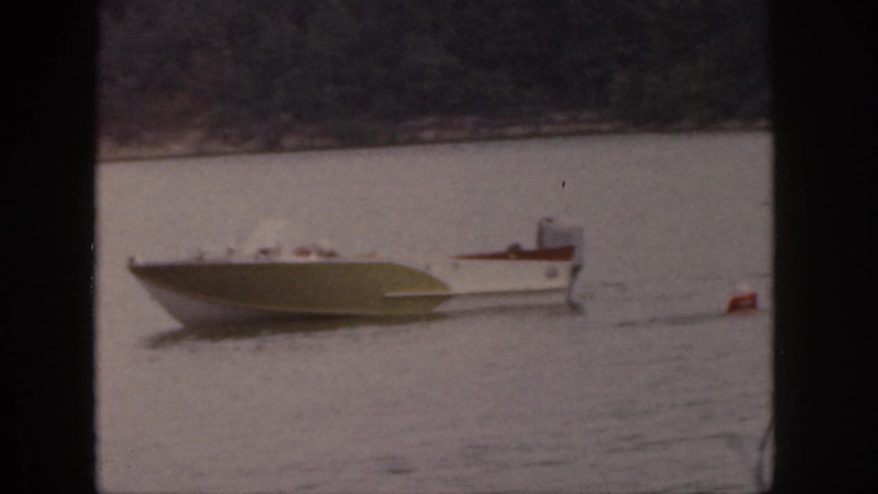 1961 Michigan home-movie frame showing a small white and green motorboat on a wooded lake with a red tow float trailing nearby.