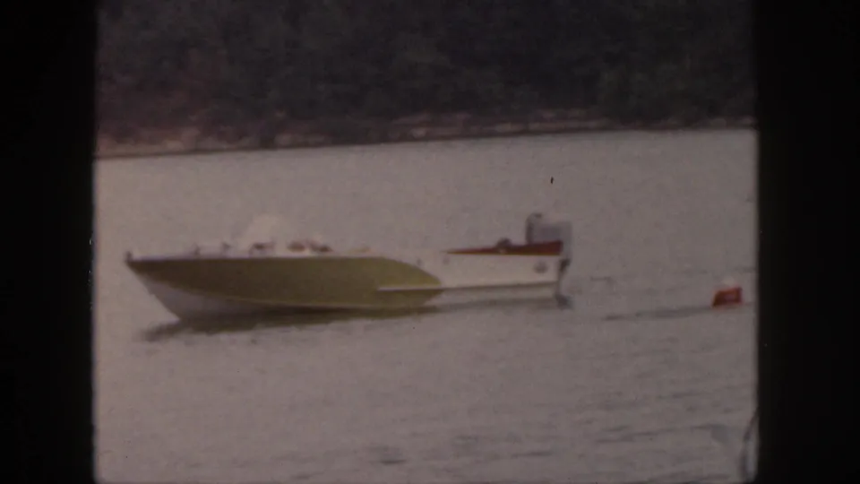 1961 Michigan home-movie frame showing a small white and green motorboat on a wooded lake with a red tow float trailing nearby.