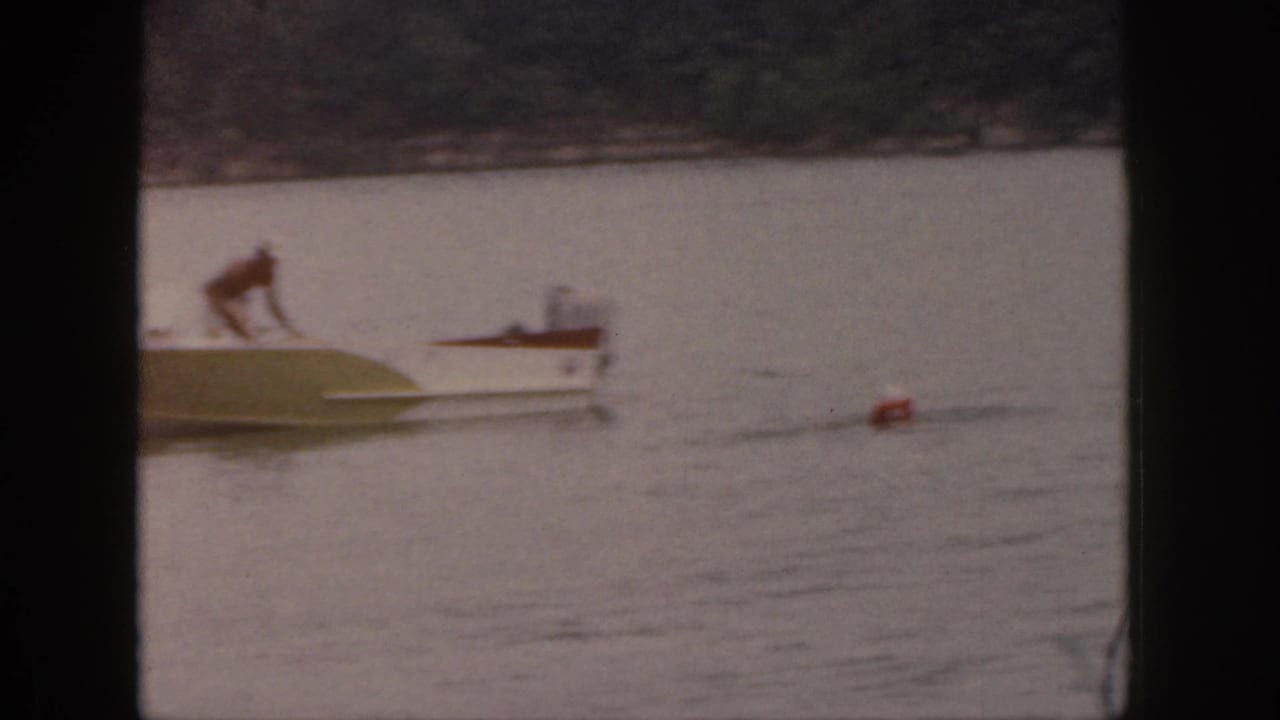 1961 Michigan 8mm frame with a person crouched on the back of a small motorboat near a trailing red float on the lake.