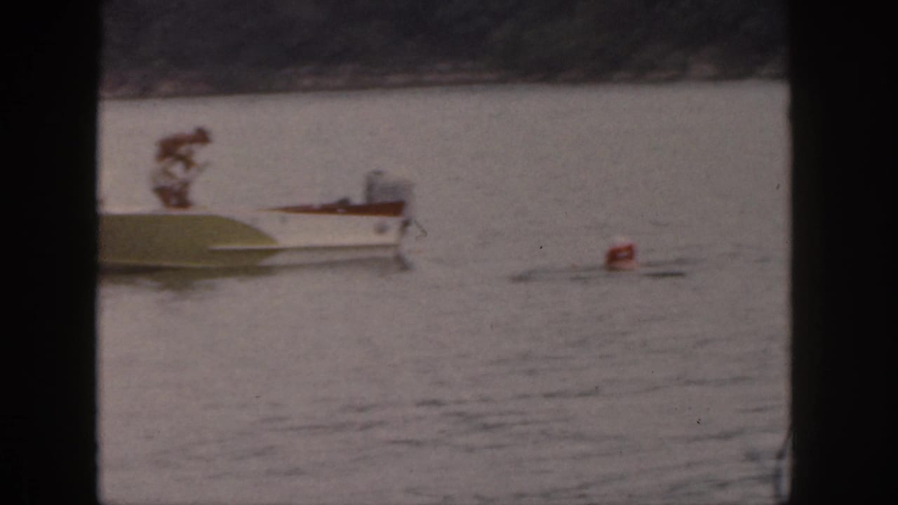 1961 Michigan home-movie frame showing a man standing on a small motorboat near a red buoy on calm lake water.