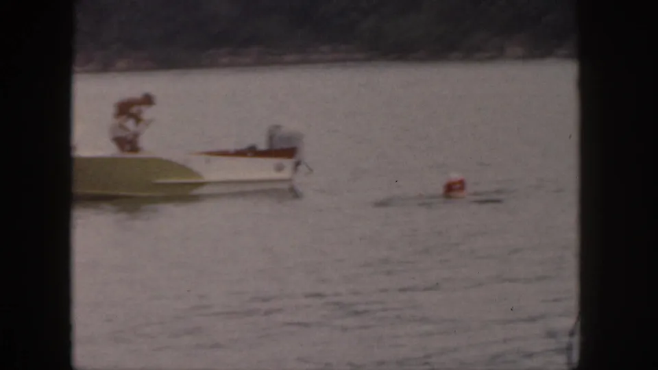 1961 Michigan home-movie frame showing a man standing on a small motorboat near a red buoy on calm lake water.