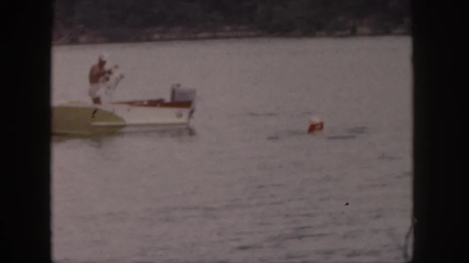 1961 Michigan lake frame with a man standing on a small motorboat as a red buoy drifts in the water nearby.